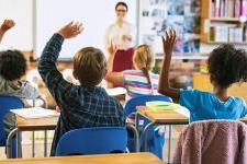 Seen from the back of a classroom, rows of young students raise their hands as their smiling teacher looks on.